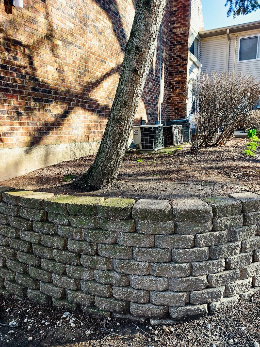 Curved block retaining wall around a mulched tree bed beside a brick house